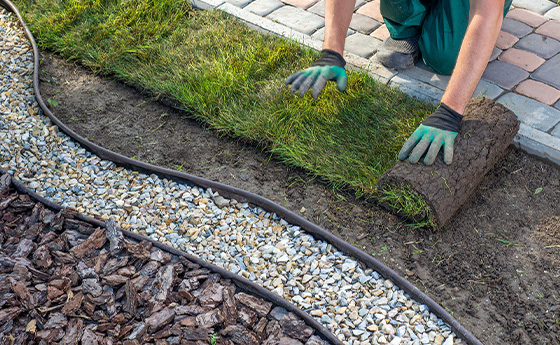 person laying sod on soil beside gravel landscaping with two types of mulch