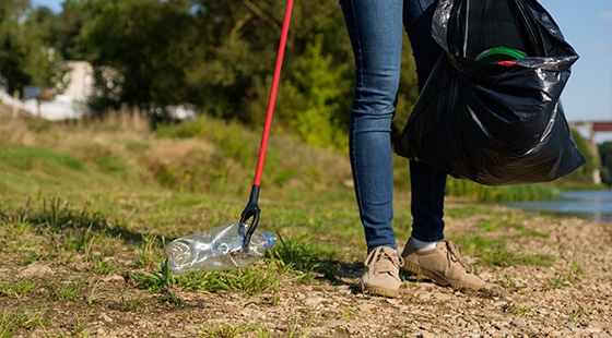 person cleaning up litter by a waterway using a grabber tool holding a black garbage bag and collecting plastic bottles three forms of pollution reduction